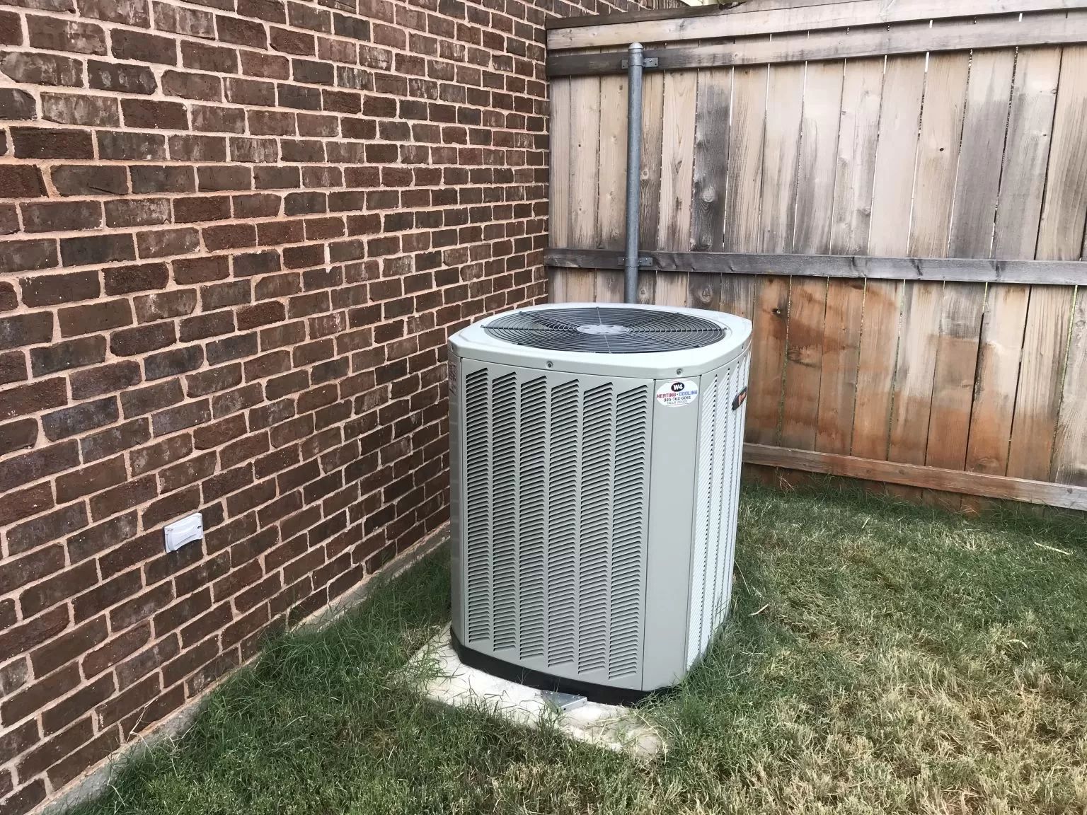 Outdoor air conditioning unit beside a brick wall and wooden fence in a small yard
