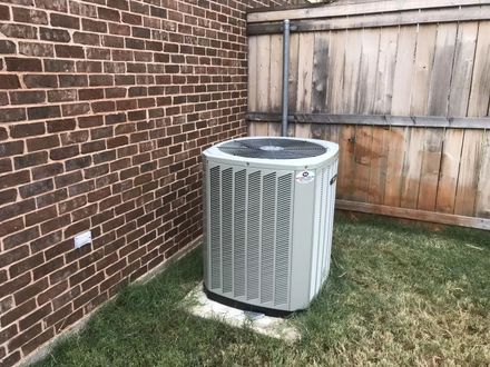 Outdoor air conditioner unit beside a brick wall and wooden fence in a grassy yard.