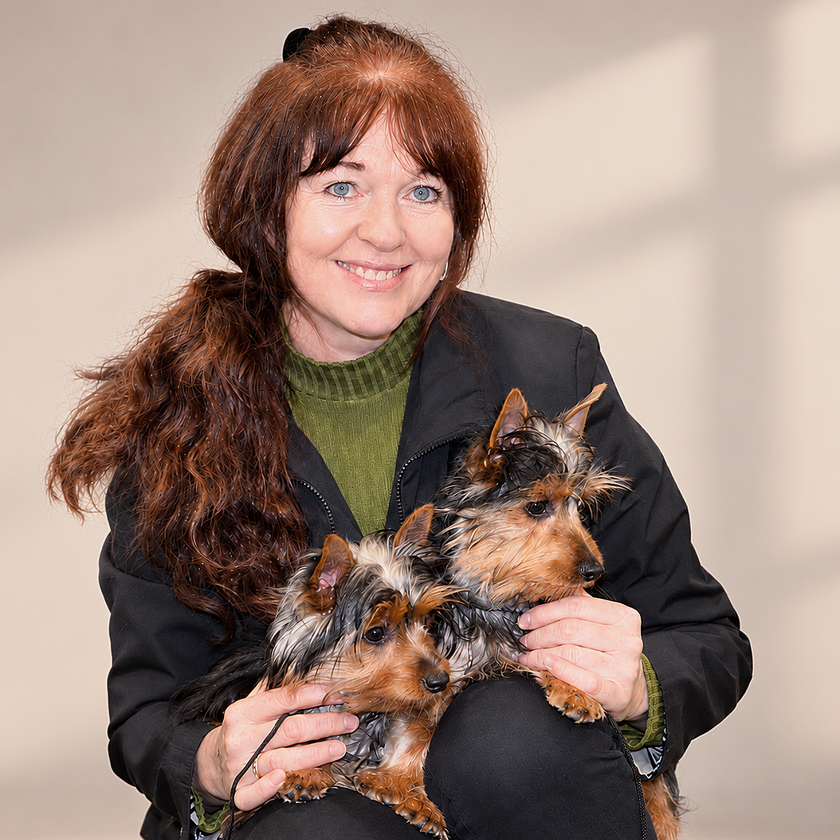 Woman holding two small dogs, smiling against a plain indoor background.