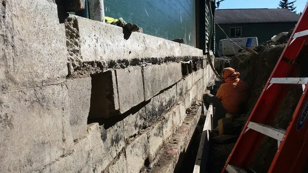 Worker repairing a cracked concrete wall beside a red ladder in bright sunlight