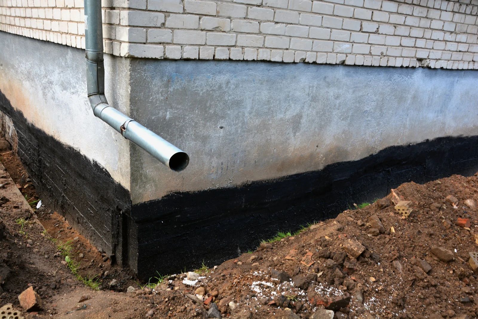 Brick building foundation with a white downspout and black waterproofing along the base.