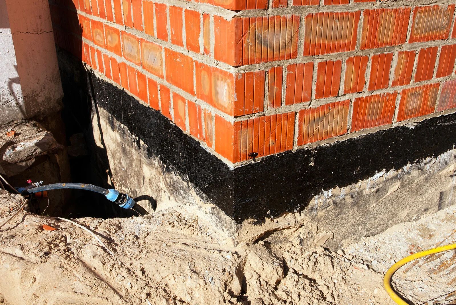 Brick building corner with black waterproofing strip at the base and exposed dirt around it