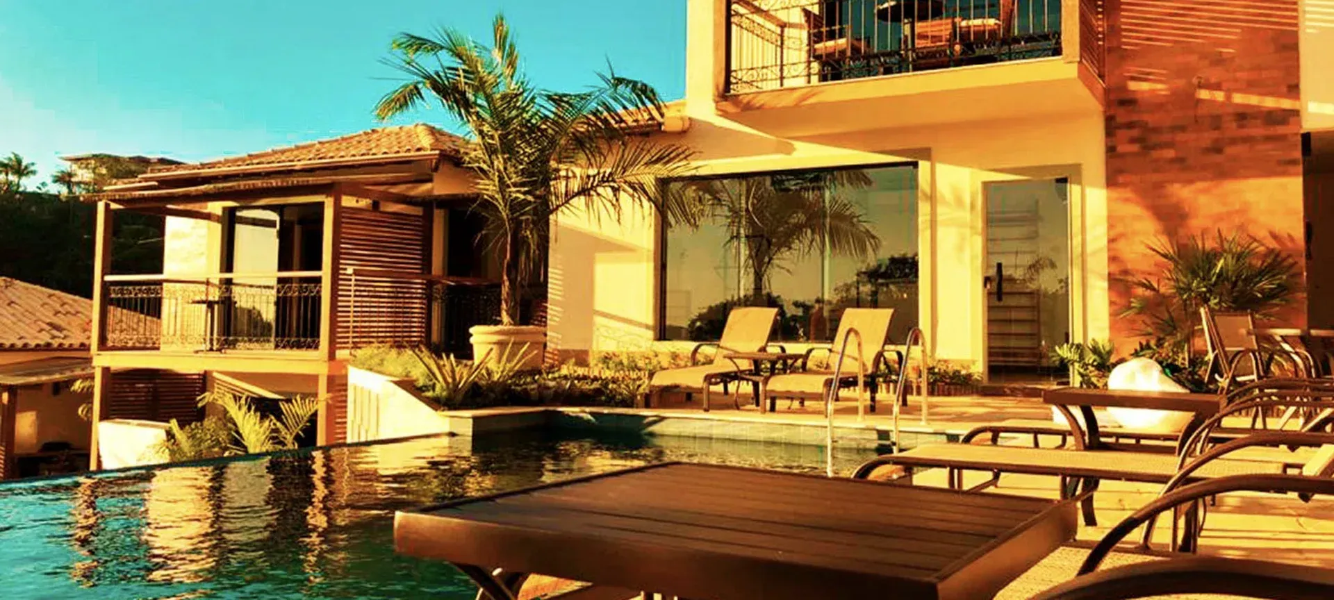 Piscina ensolarada do resort com espreguiçadeiras e uma villa moderna, vista da água.