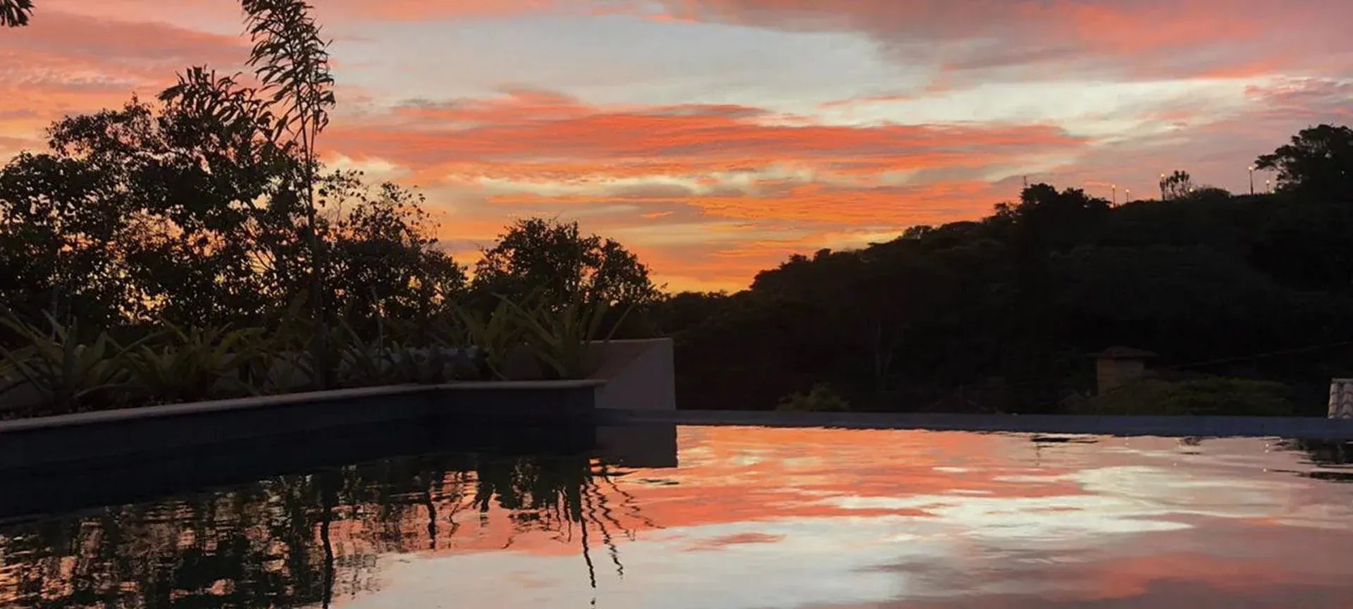 Céu ao pôr do sol refletido em uma piscina tranquila ao lado de árvores silhuetadas e uma encosta escura.