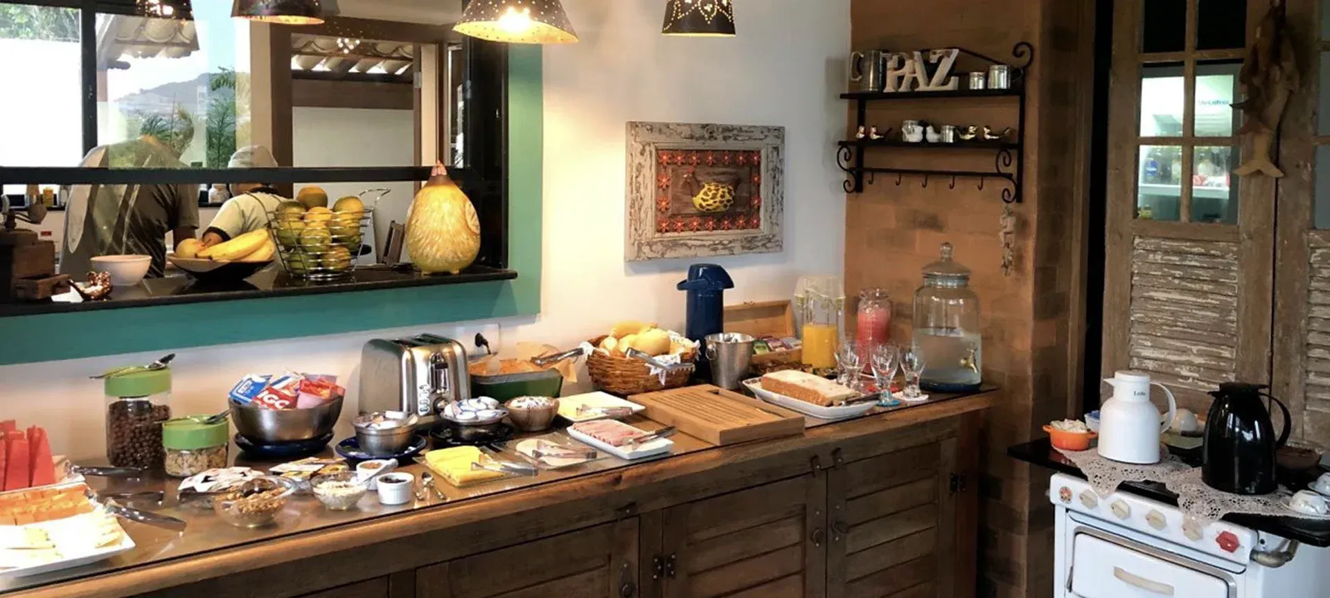 Mesa de buffet rústica na cozinha, com doces, frutas e café, sob uma janela e luminárias pendentes.