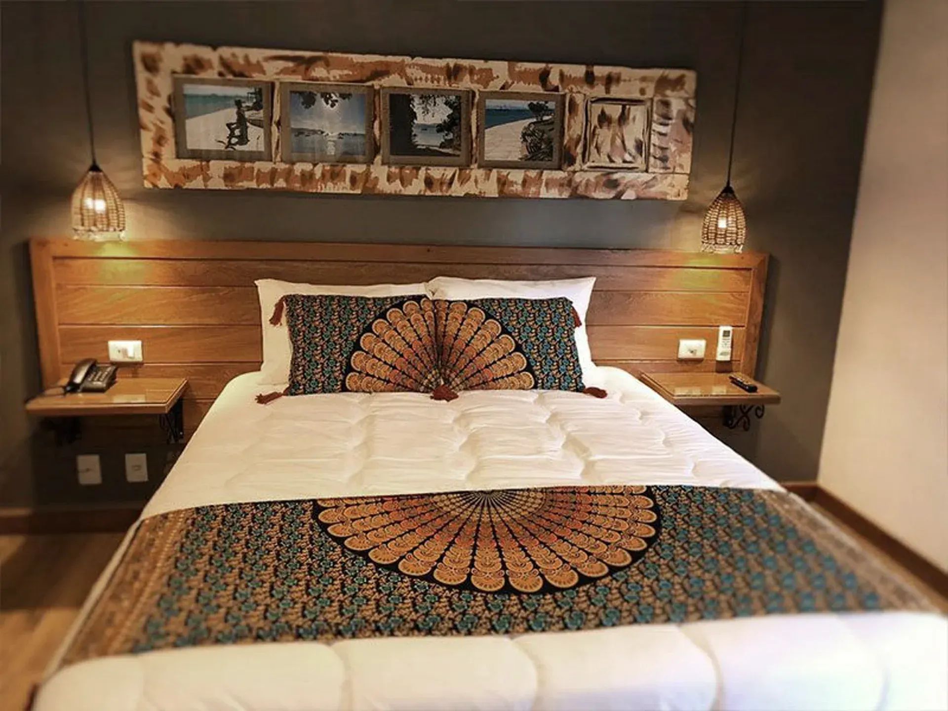 Quarto com cama arrumada, cabeceira com painéis de madeira, luminárias aconchegantes e obras de arte decorativas na parede.