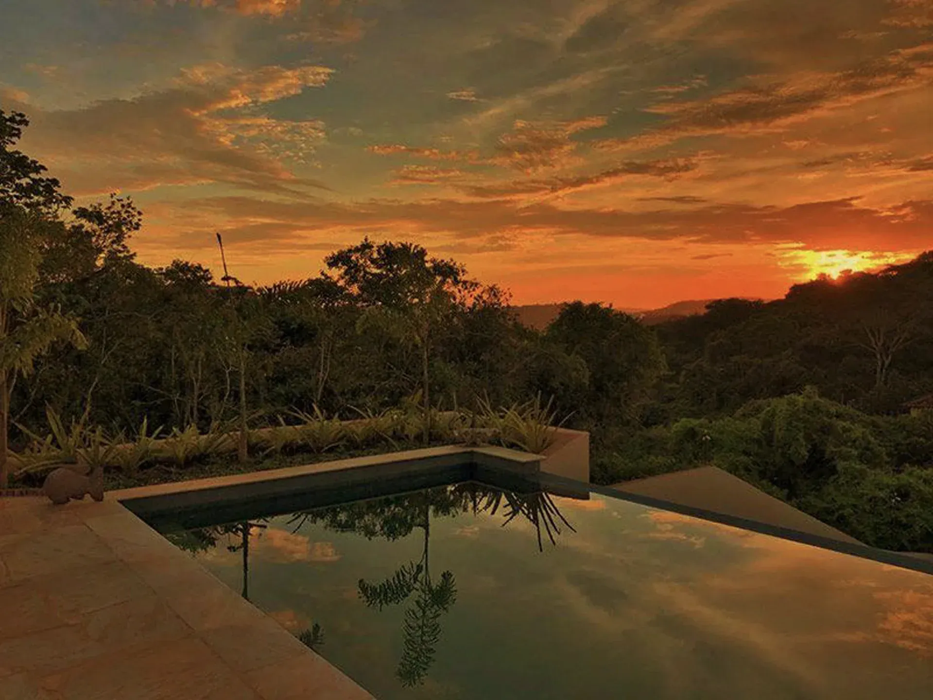 Pôr do sol sobre uma piscina na encosta de uma colina tropical, com o céu alaranjado refletido na água calma.