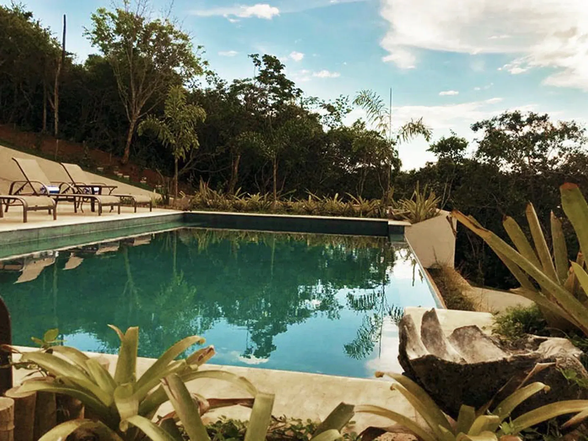 Piscina de resort tropical com água turquesa, espreguiçadeiras e árvores exuberantes sob um céu azul.