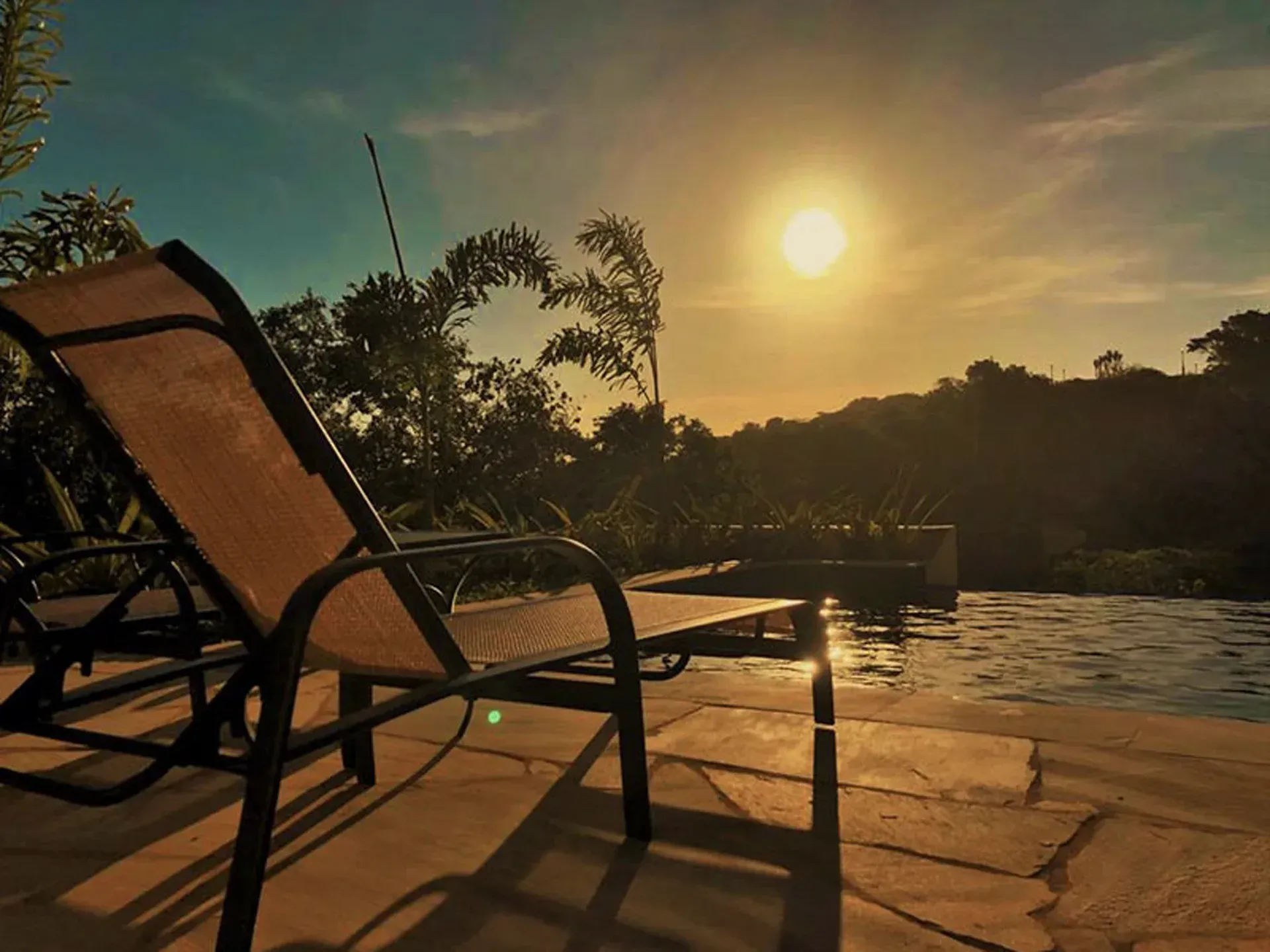 Cadeiras de pátio ensolaradas à beira da piscina ao pôr do sol, com árvores em silhueta ao fundo.