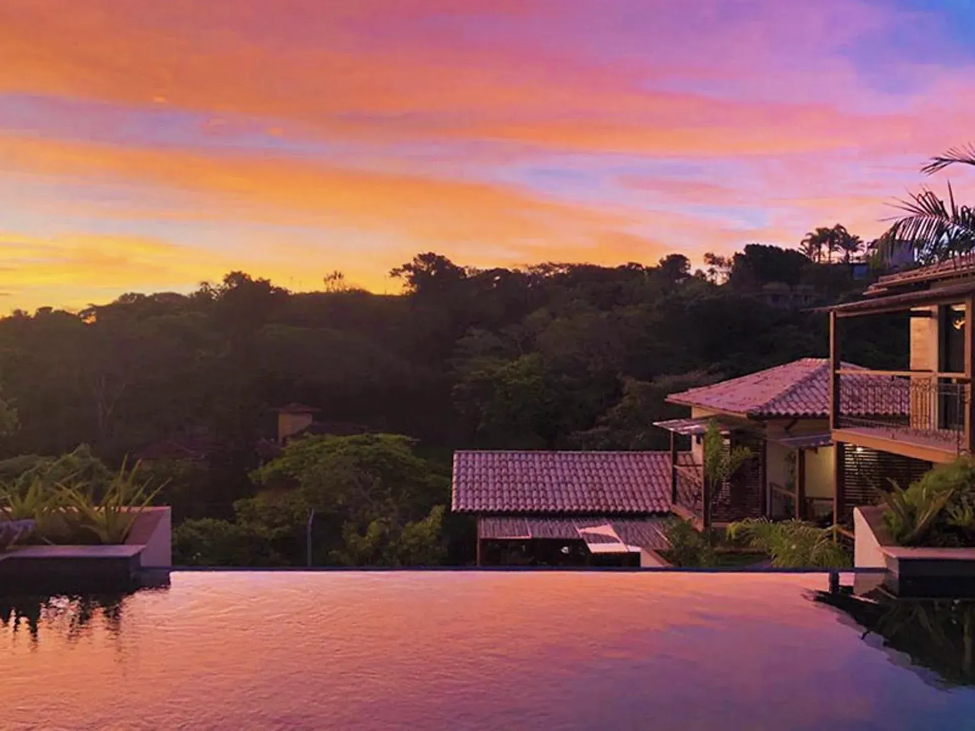Piscina infinita ao pôr do sol com vista para colinas verdejantes e vilas com telhados de azulejo, com um céu rosa.