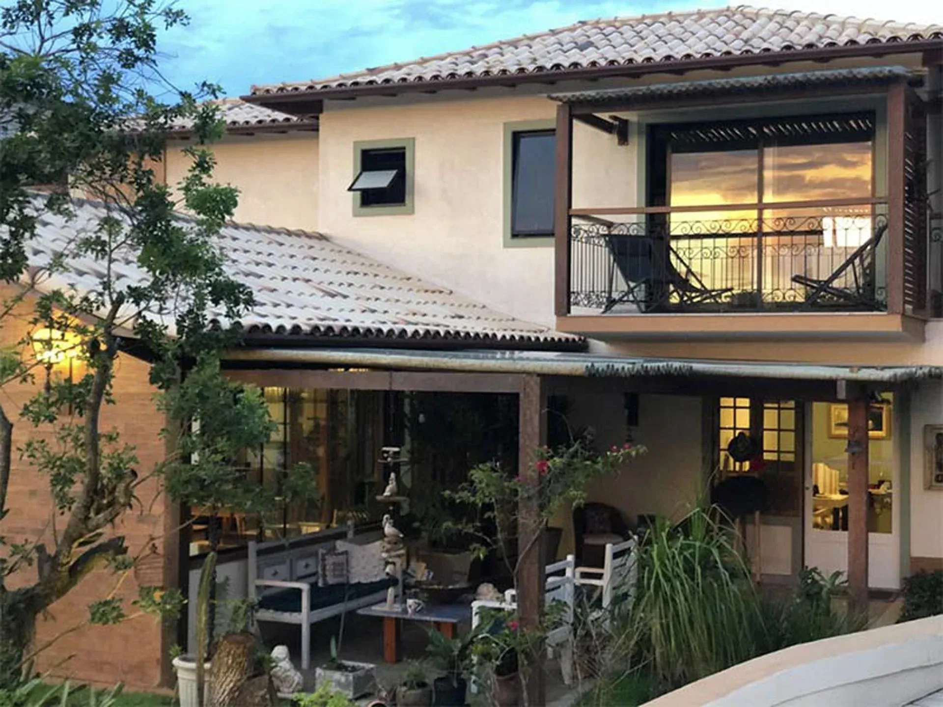 Casa bege de dois andares com telhado de telha, pátio coberto e luzes aconchegantes que brilham ao entardecer.