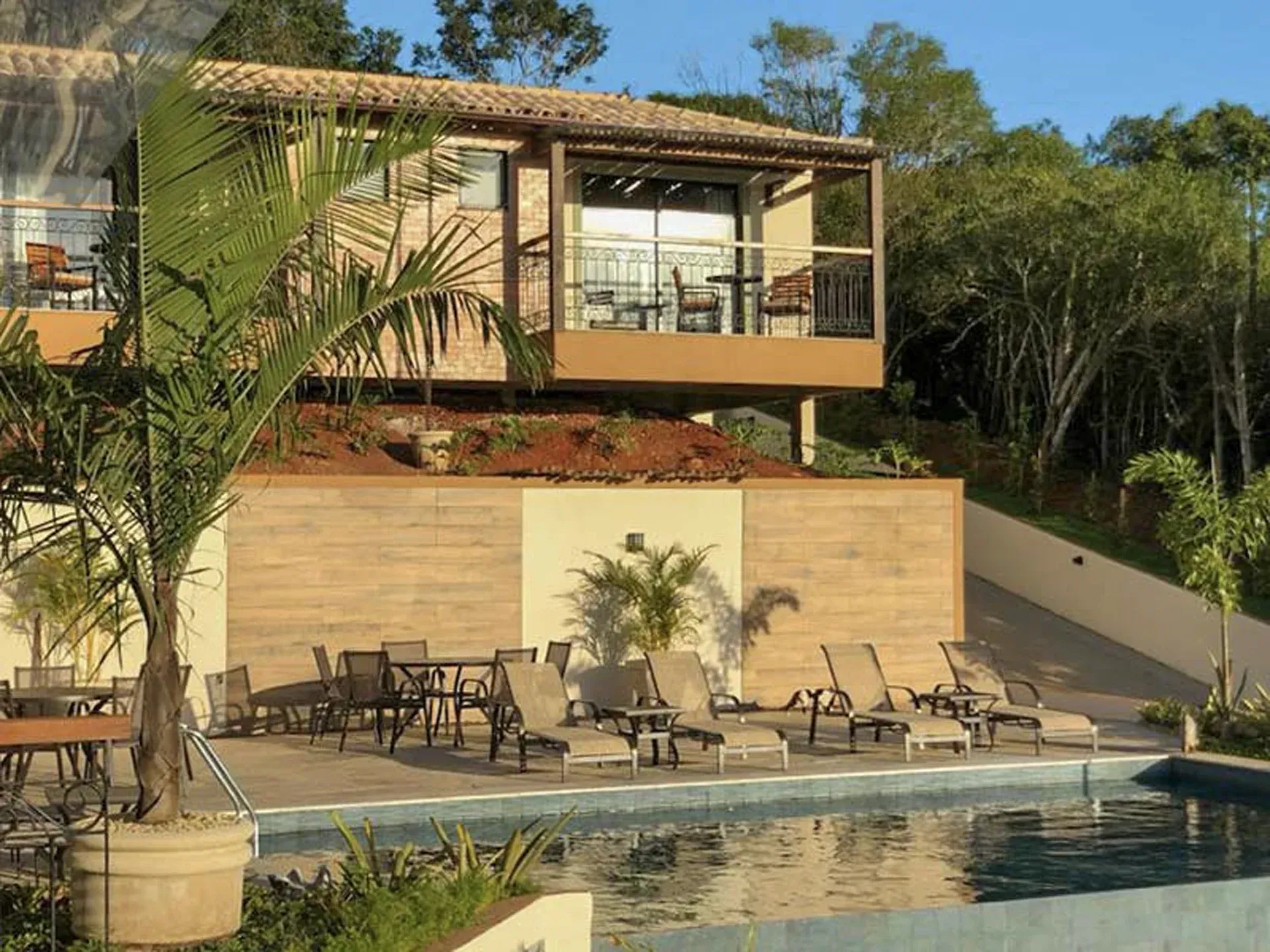 Piscina moderna de resort com espreguiçadeiras, palmeiras e um edifício contemporâneo em uma encosta ensolarada.