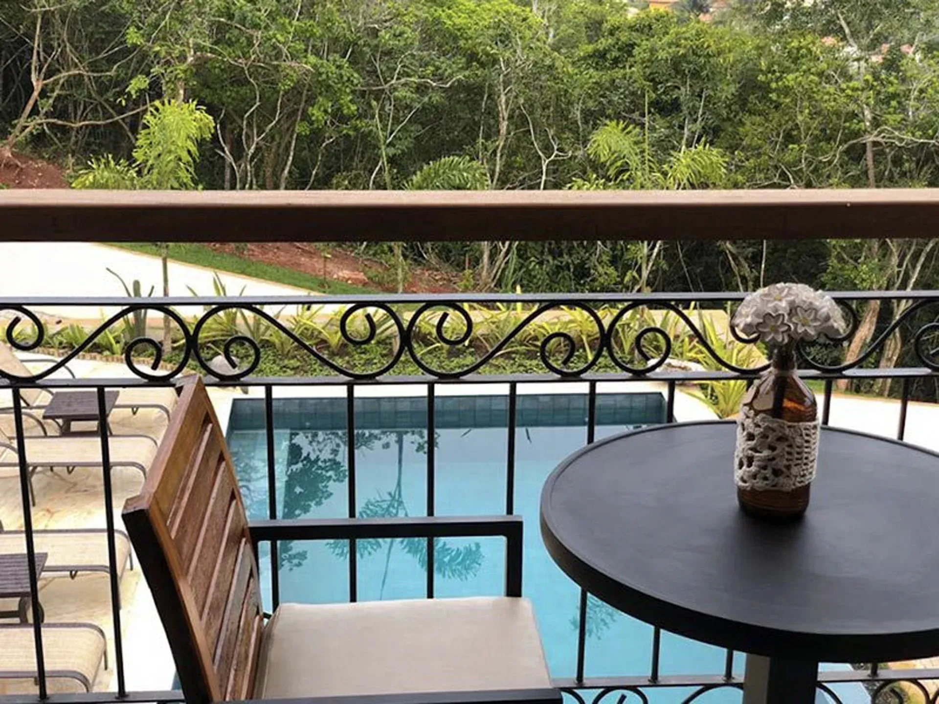 Varanda com mesa, cadeira e vaso pretos, com vista para uma piscina turquesa e vegetação exuberante.