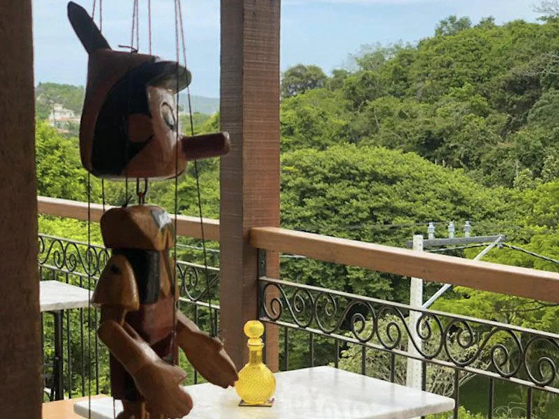 Figura esculpida em madeira pendurada em uma sacada com colinas verdes e balaustrada ao fundo.