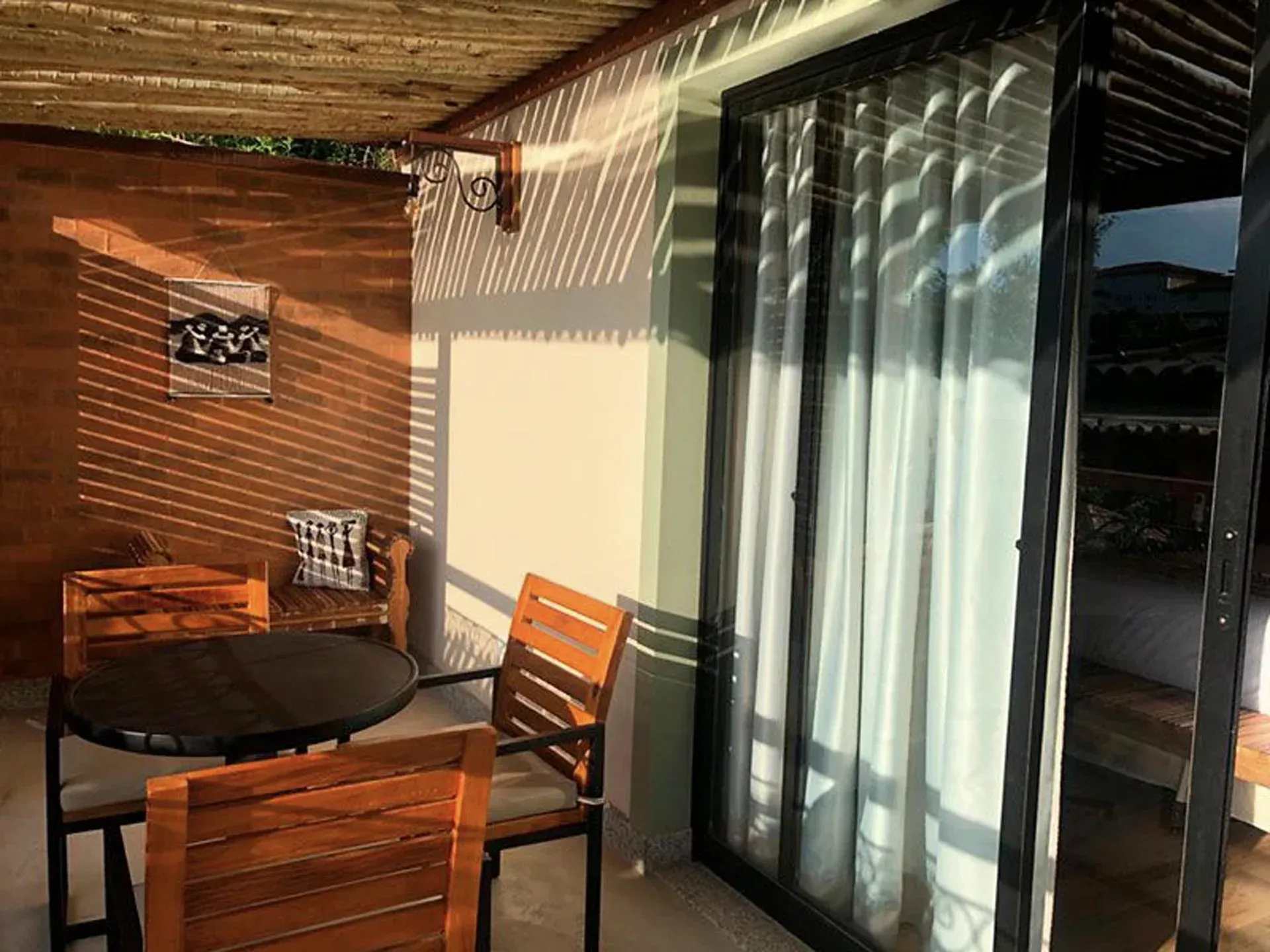Área de estar externa com mesa e cadeiras de madeira ao lado de uma porta de vidro deslizante e cortinas translúcidas.