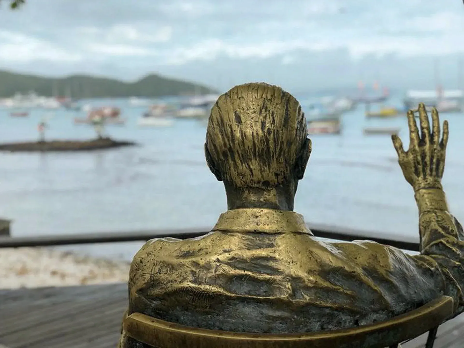 Estátua de bronze vista de costas, com a mão erguida ao lado de um porto com barcos e colinas ao longe.