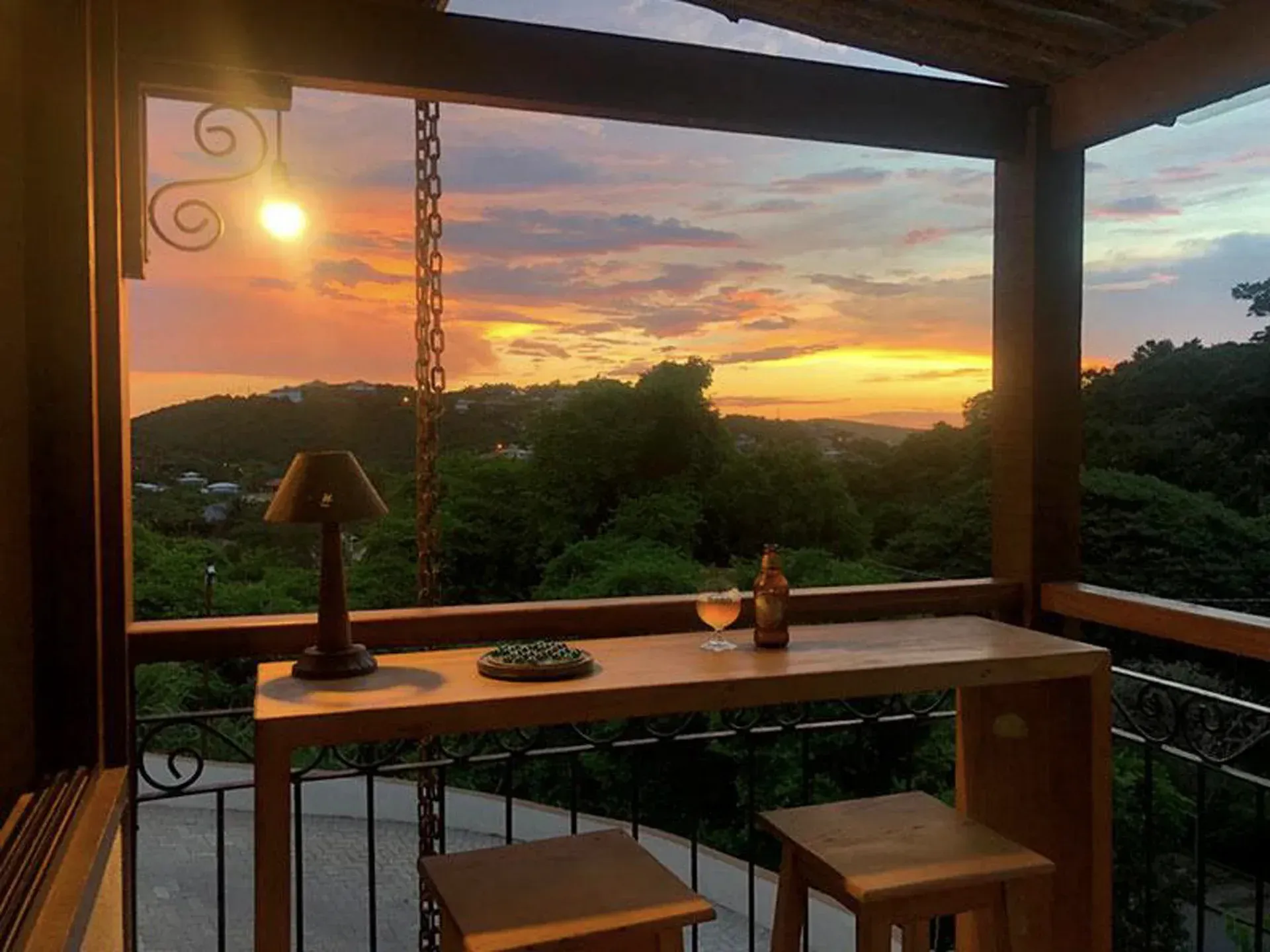 Vista do pôr do sol de uma varanda com mesa de madeira, cadeiras e um candeeiro suspenso, com vista.