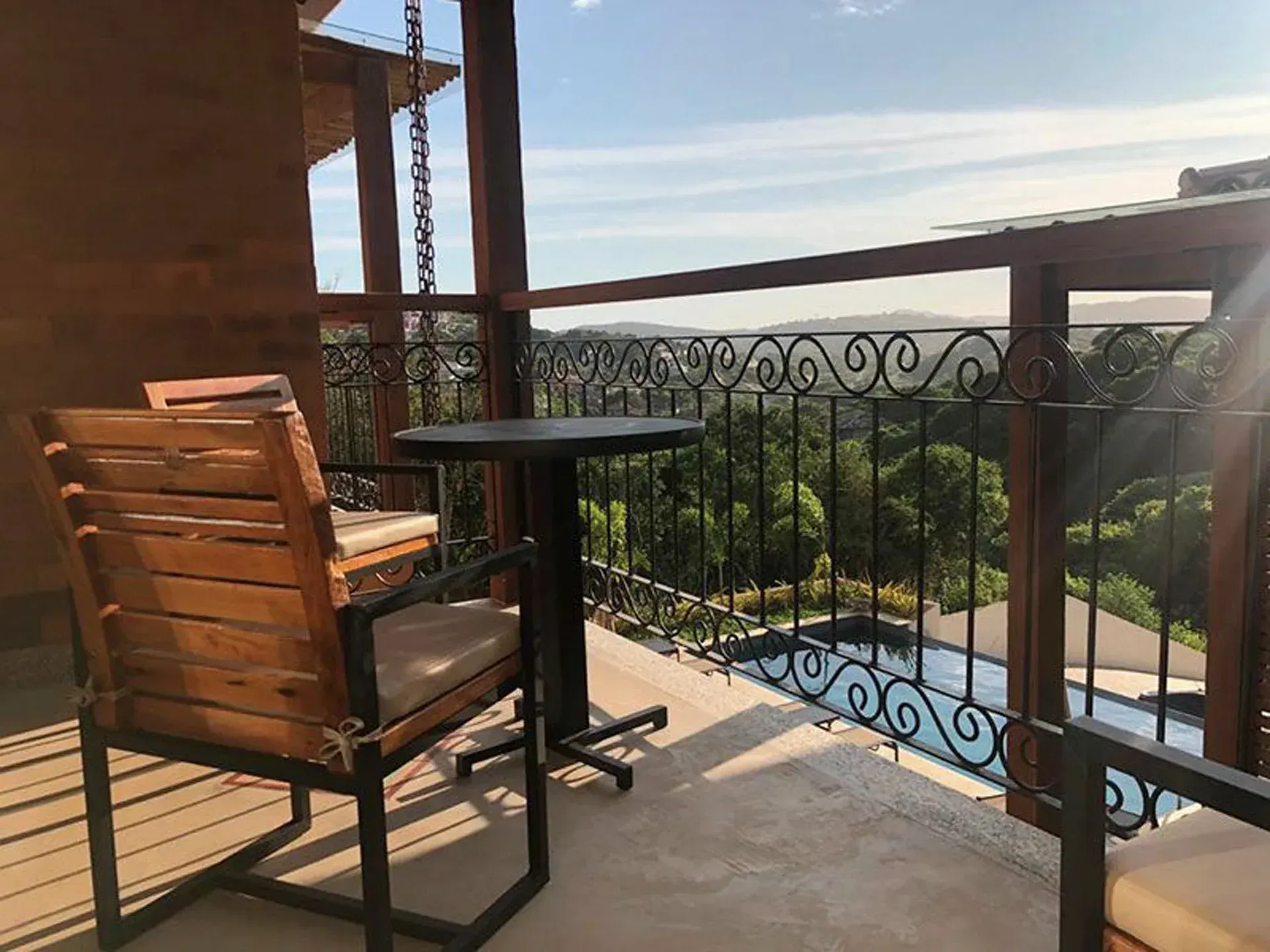 Varanda com cadeira de madeira, mesa redonda e balaustrada ornamentada com vista para a piscina e a vegetação ao pôr do sol.