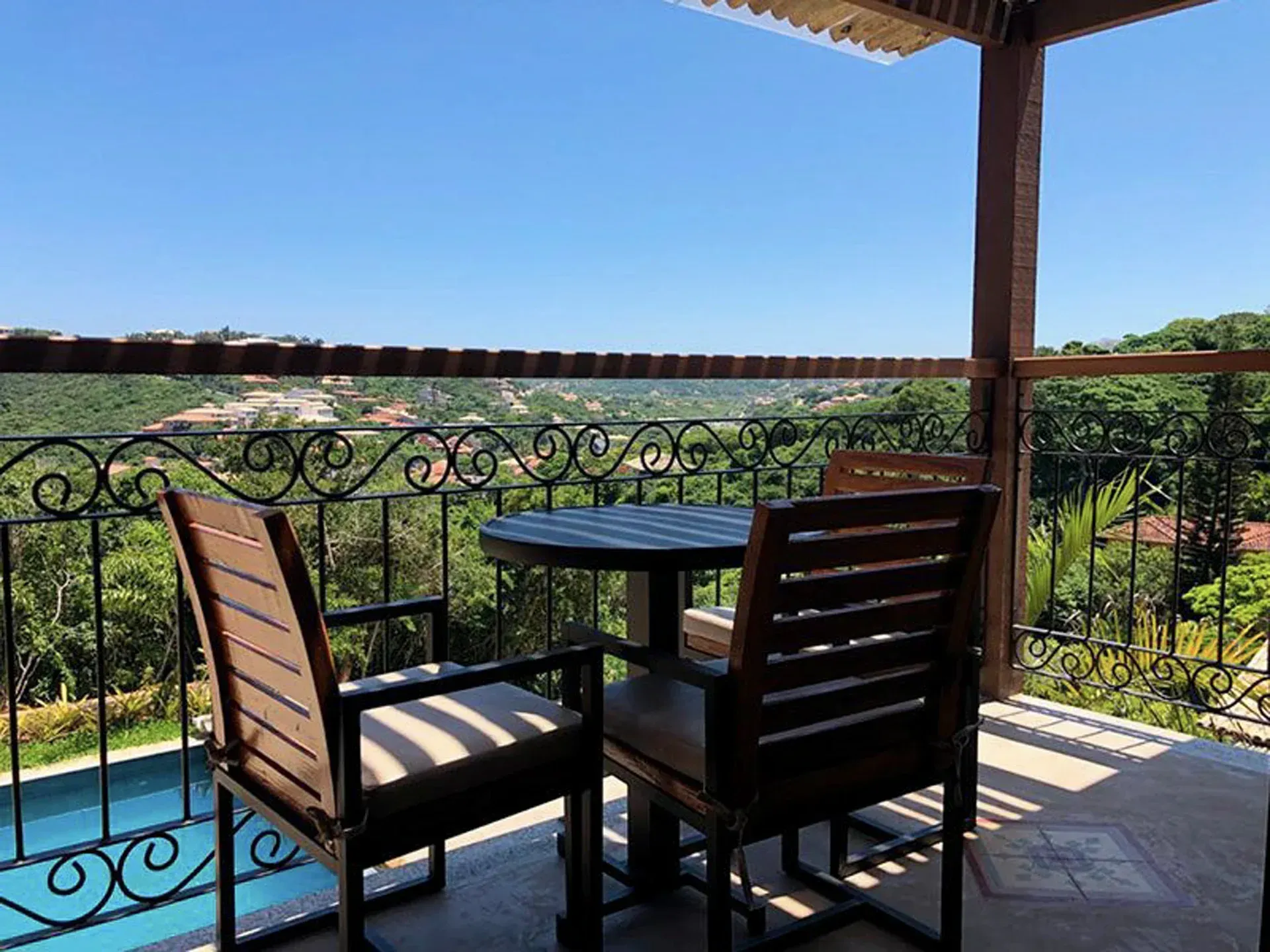 Mesa e cadeiras de pátio sob uma pérgola sombreada com vista para uma paisagem verdejante e água azul.