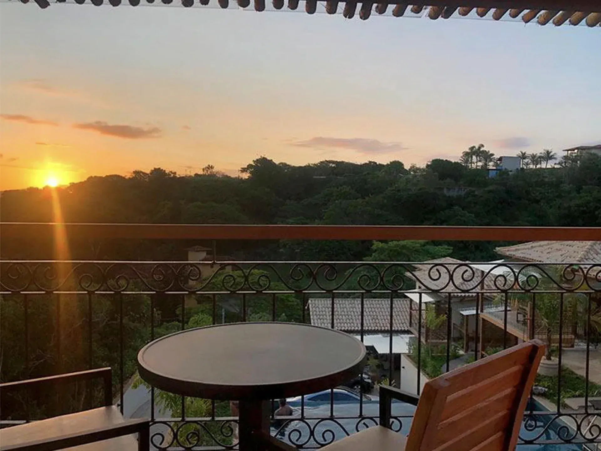 Vista do pôr do sol de uma varanda com mesa e cadeira, com vista para árvores e telhados.