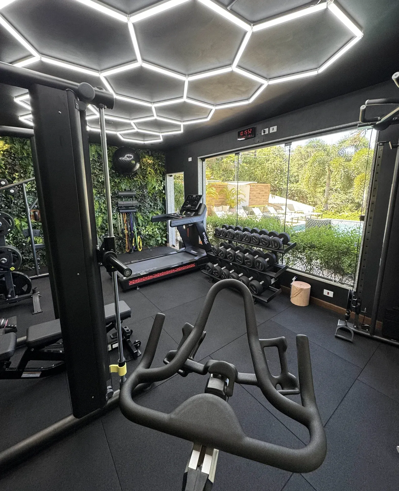 Academia moderna com luminárias hexagonais no teto, bicicletas ergométricas, halteres e grandes janelas com vista.