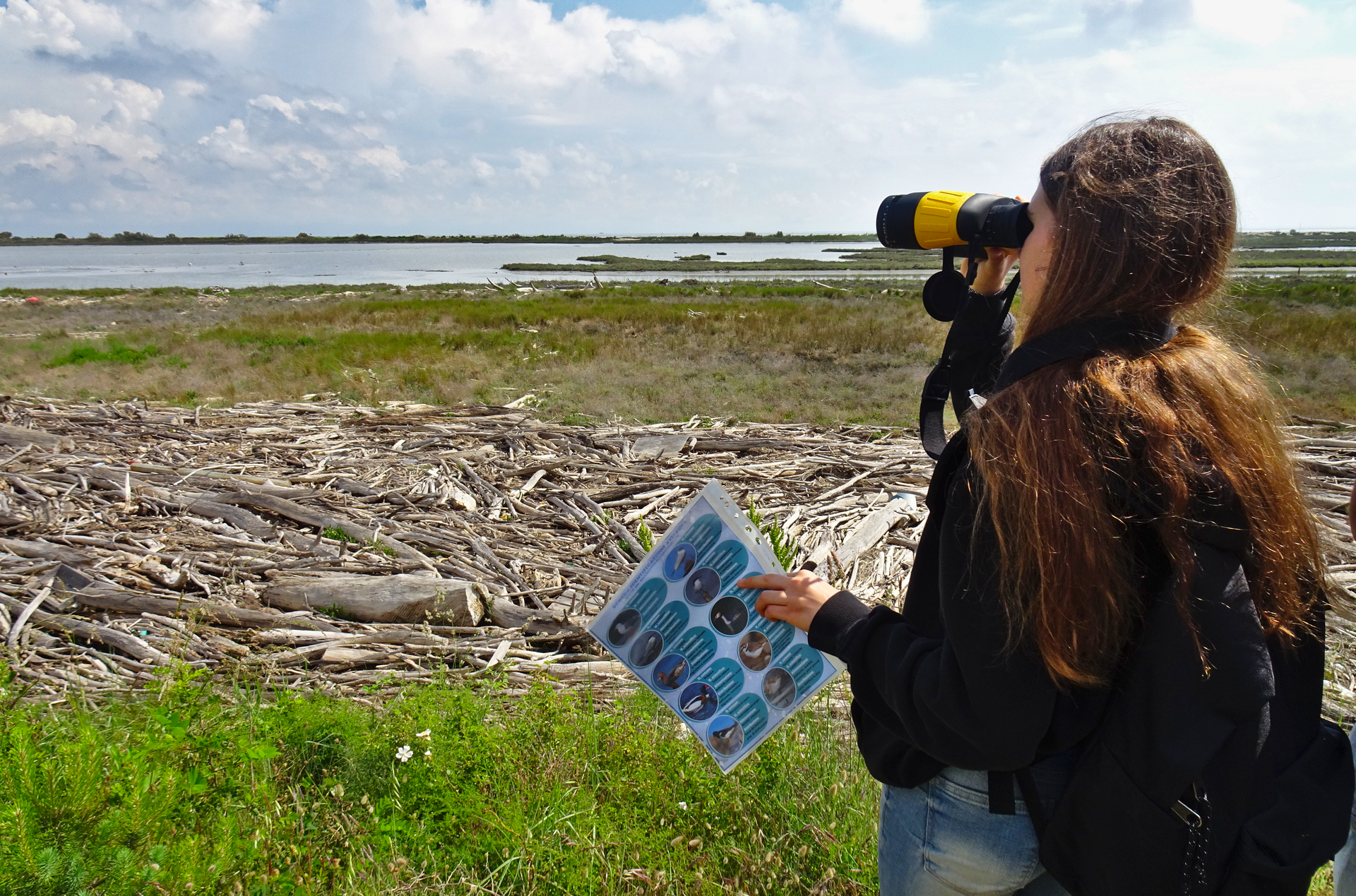 Studentessa che utilizza un binocolo in riva al mare, con in mano una guida naturalistica in una area naturale.