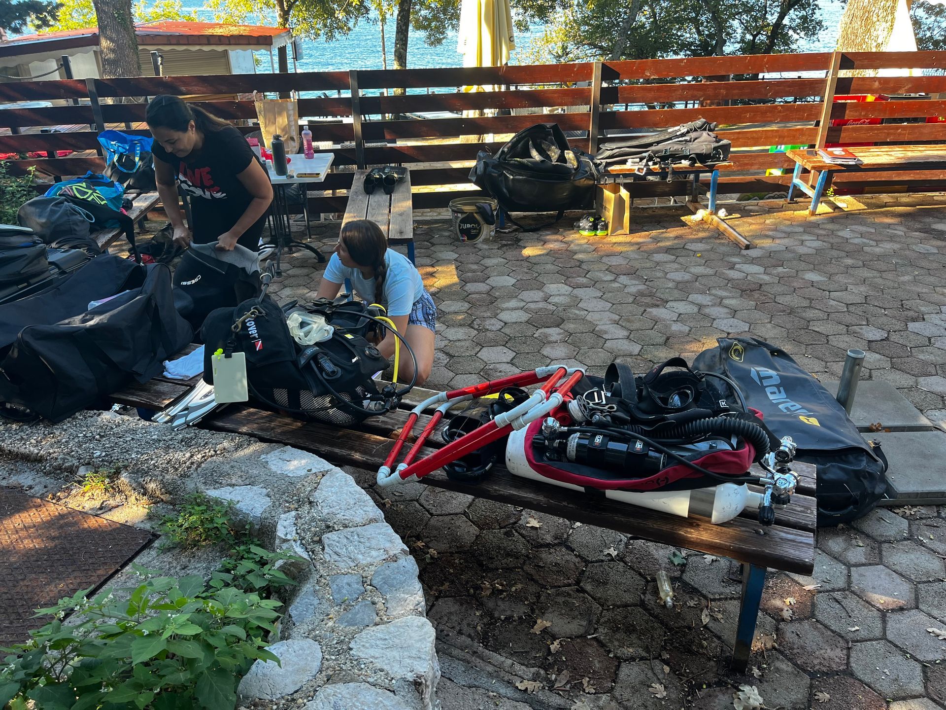 Biciclette e attrezzatura disposte su una panchina del parco, con due persone che sistemano gli oggetti su un sentiero di pietra.