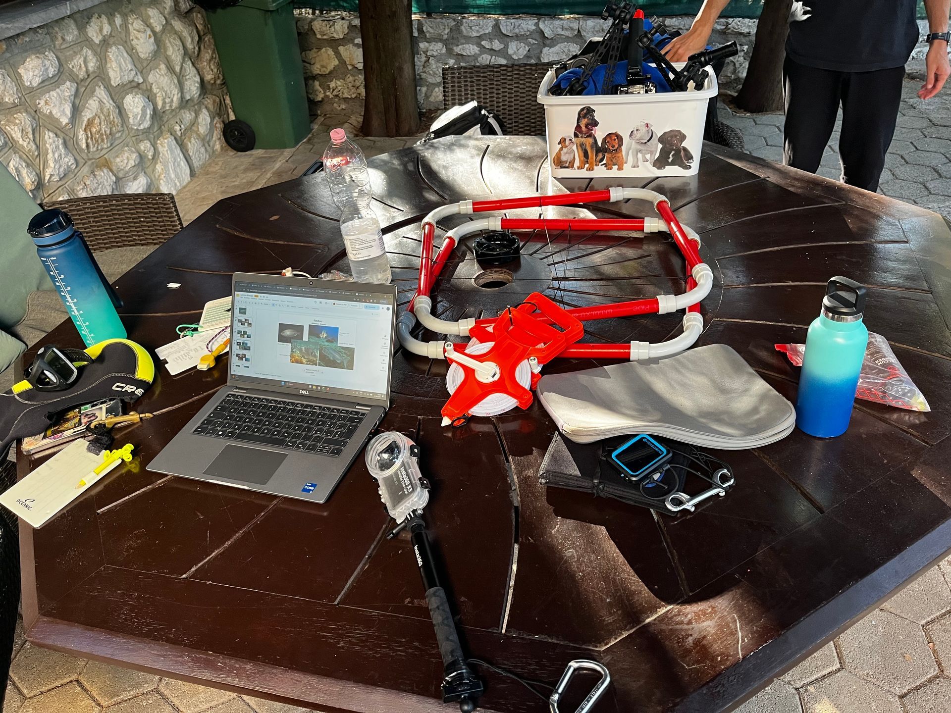 Tavolo con un computer portatile, componenti di un drone, attrezzi, un telefono e una bottiglia d’acqua in un ambiente all’aperto