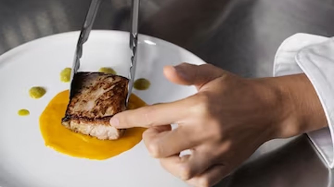 Chef plating seared fish with tongs, placing it over a yellow sauce on a white plate.