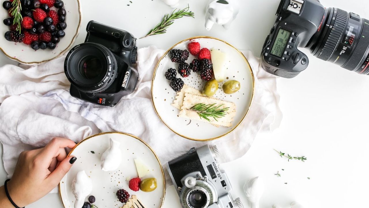 Plates of fruit and cheese with cameras on a white surface, with a hand reaching for a plate.