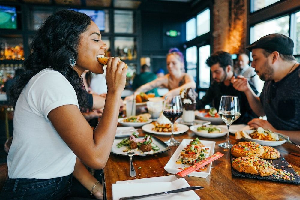People eating food at a restaurant table. One person is taking a bite. Various dishes and drinks are on the table.