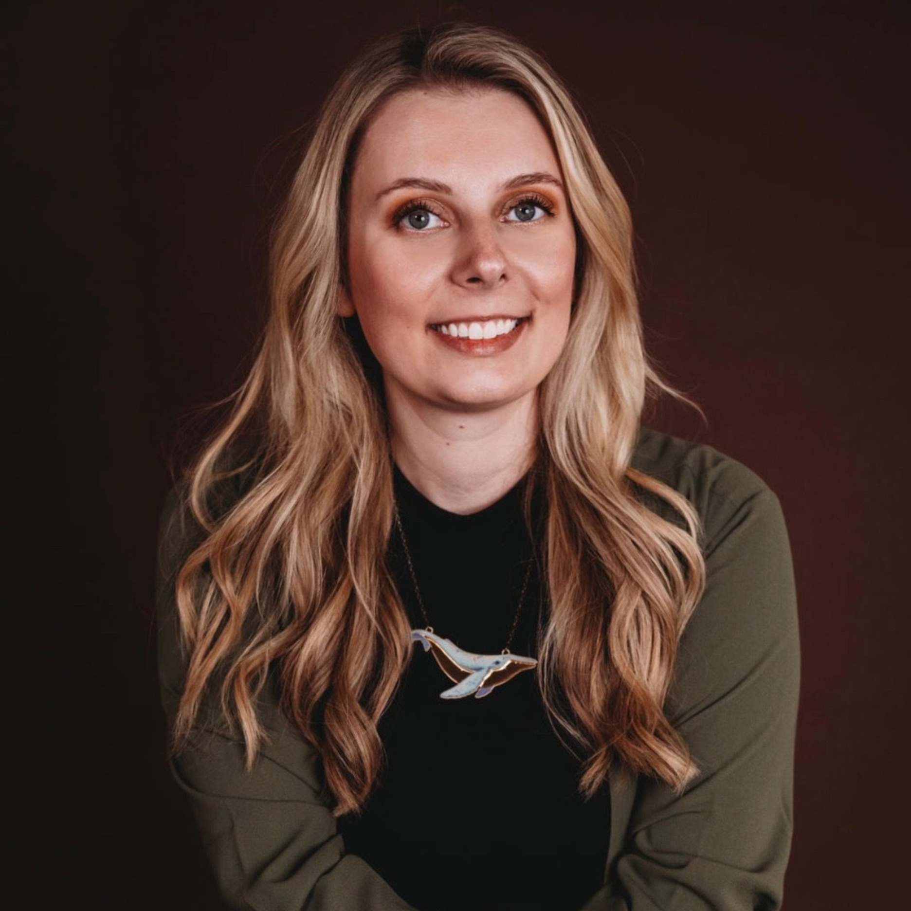 Smiling person with long blonde hair in a dark top and olive jacket against a dark background