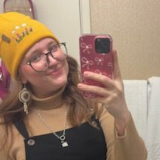 Person taking a mirror selfie in a bathroom, wearing a yellow beanie and glasses, smiling.