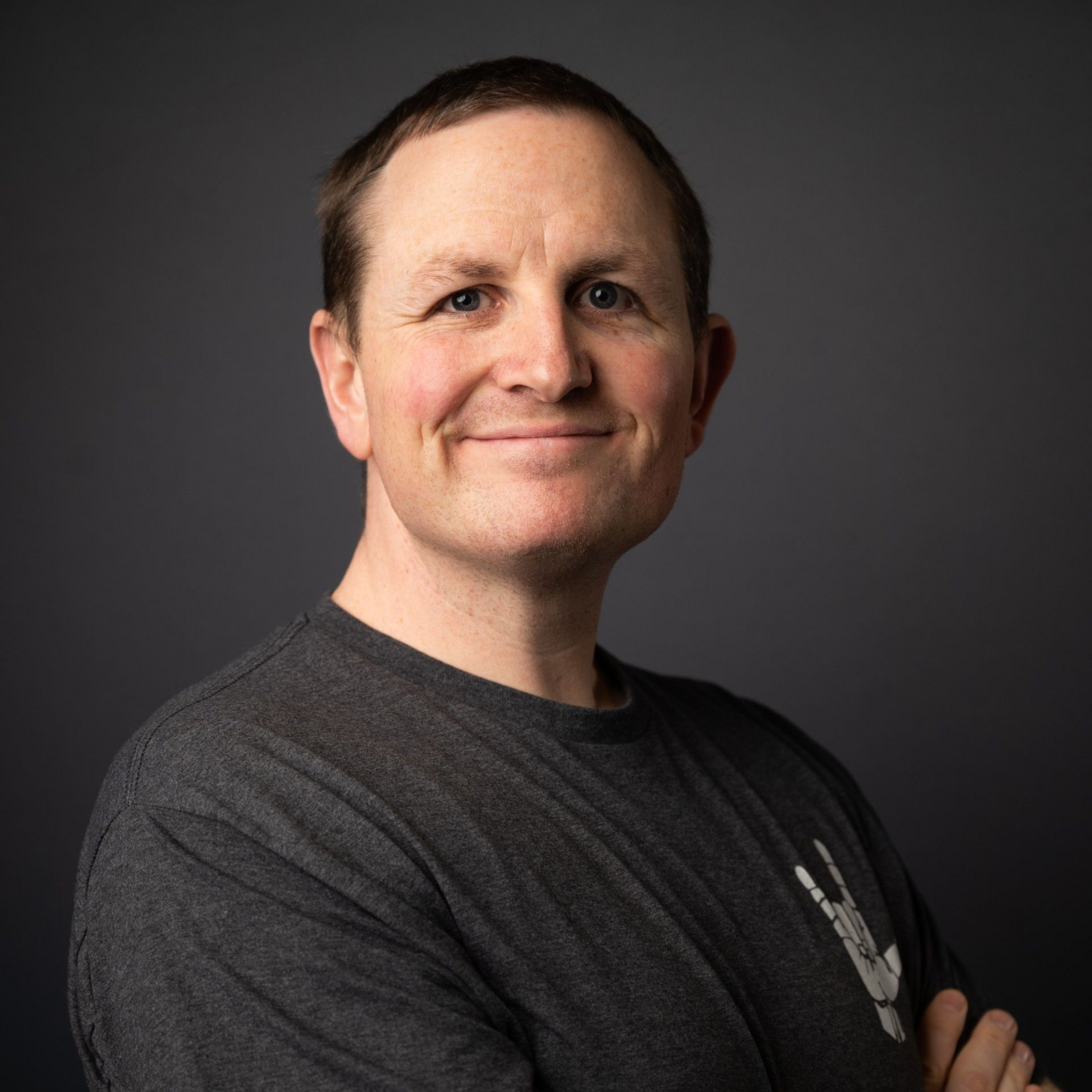 Smiling person in a dark gray shirt with arms crossed against a dark gray background