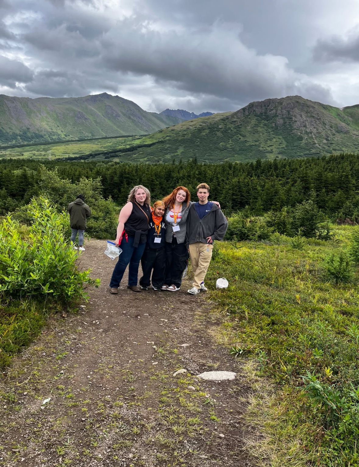 Four people posing on a gravel trail with green mountains and cloudy sky in the background