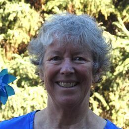 Smiling person with short gray hair in a blue shirt, standing outdoors with greenery behind them