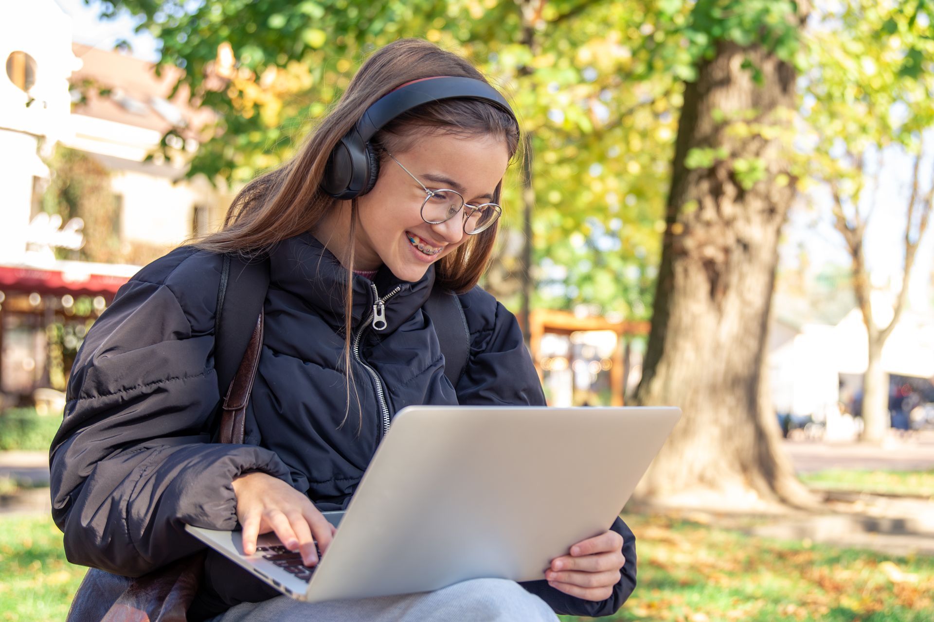 Person wearing headphones and a black jacket using a laptop outdoors in a sunny park