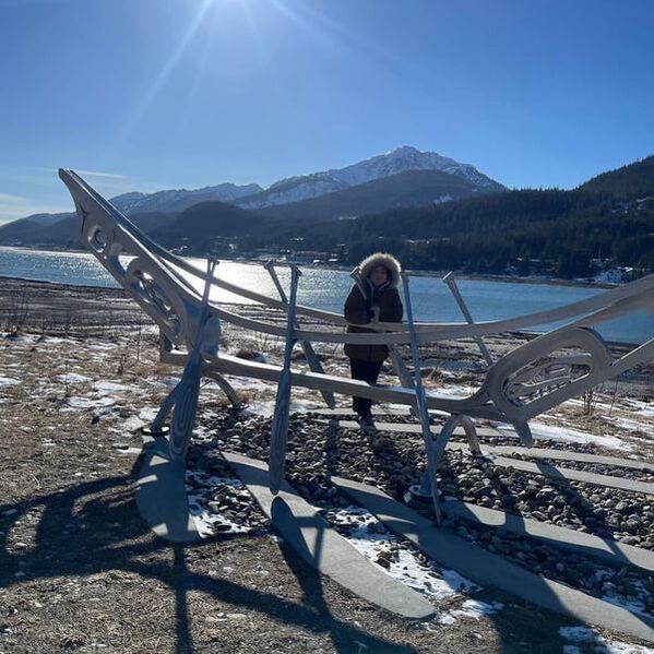 Person in a winter coat stands by a metal sculpture on a snowy lakeshore with mountains and blue sky.