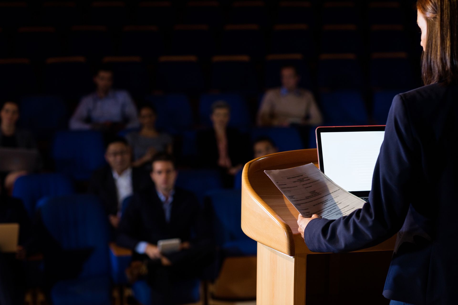 Speaker at podium addressing a dimly lit audience in a conference room