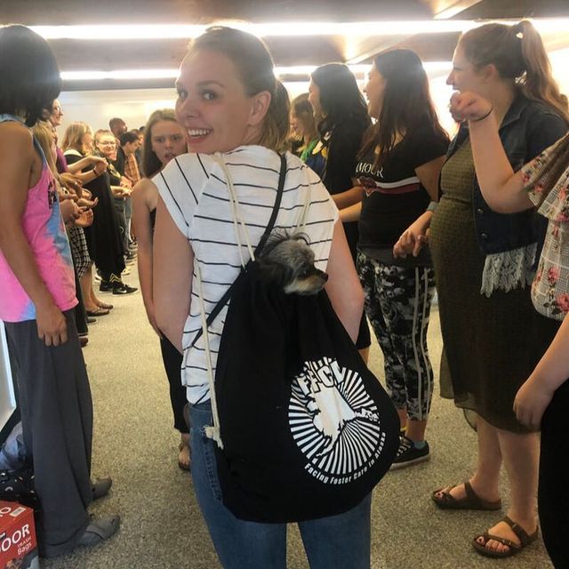 Woman in striped top smiles over her shoulder in a crowded indoor hallway, wearing a black drawstring backpack.