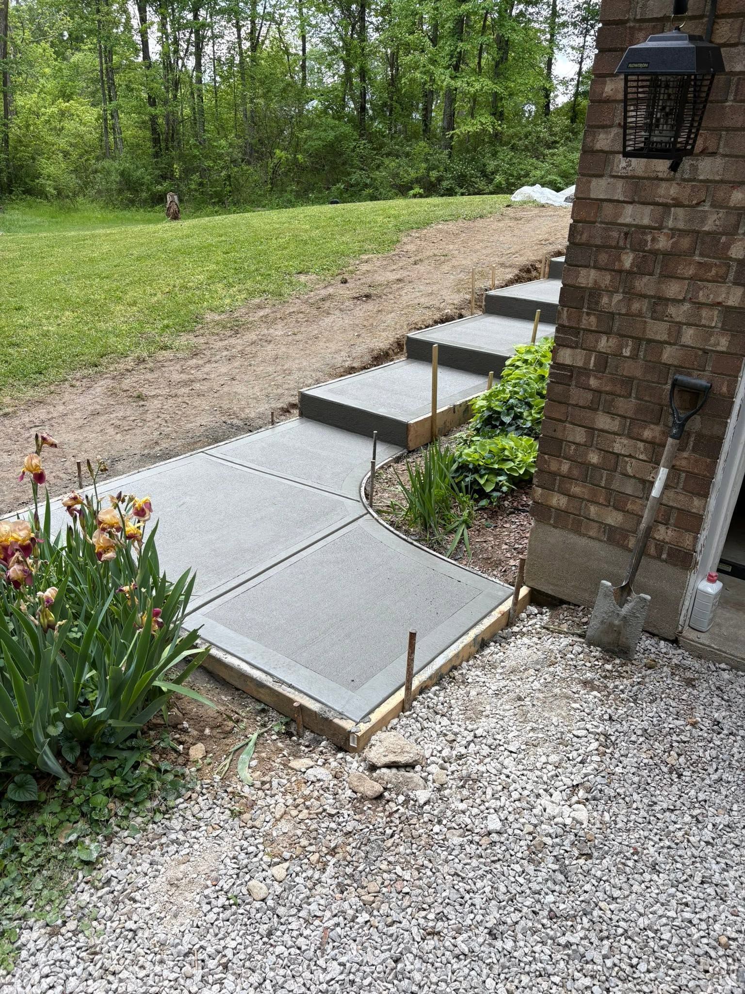 Concrete garden steps beside a brick house, leading up a gravel slope with green plants and trees beyond