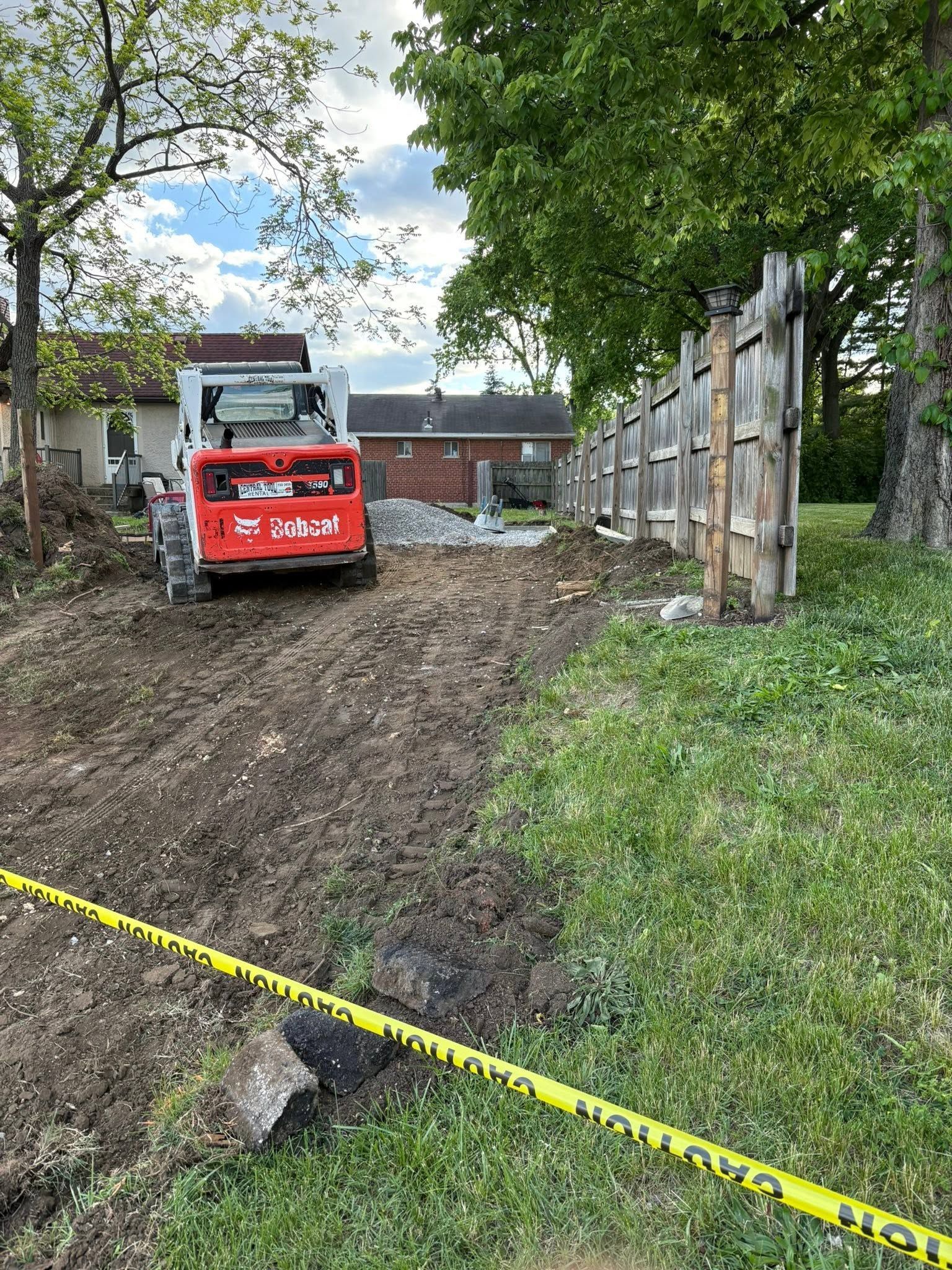 Red excavator on muddy lot beside fence, with yellow caution tape in foreground.