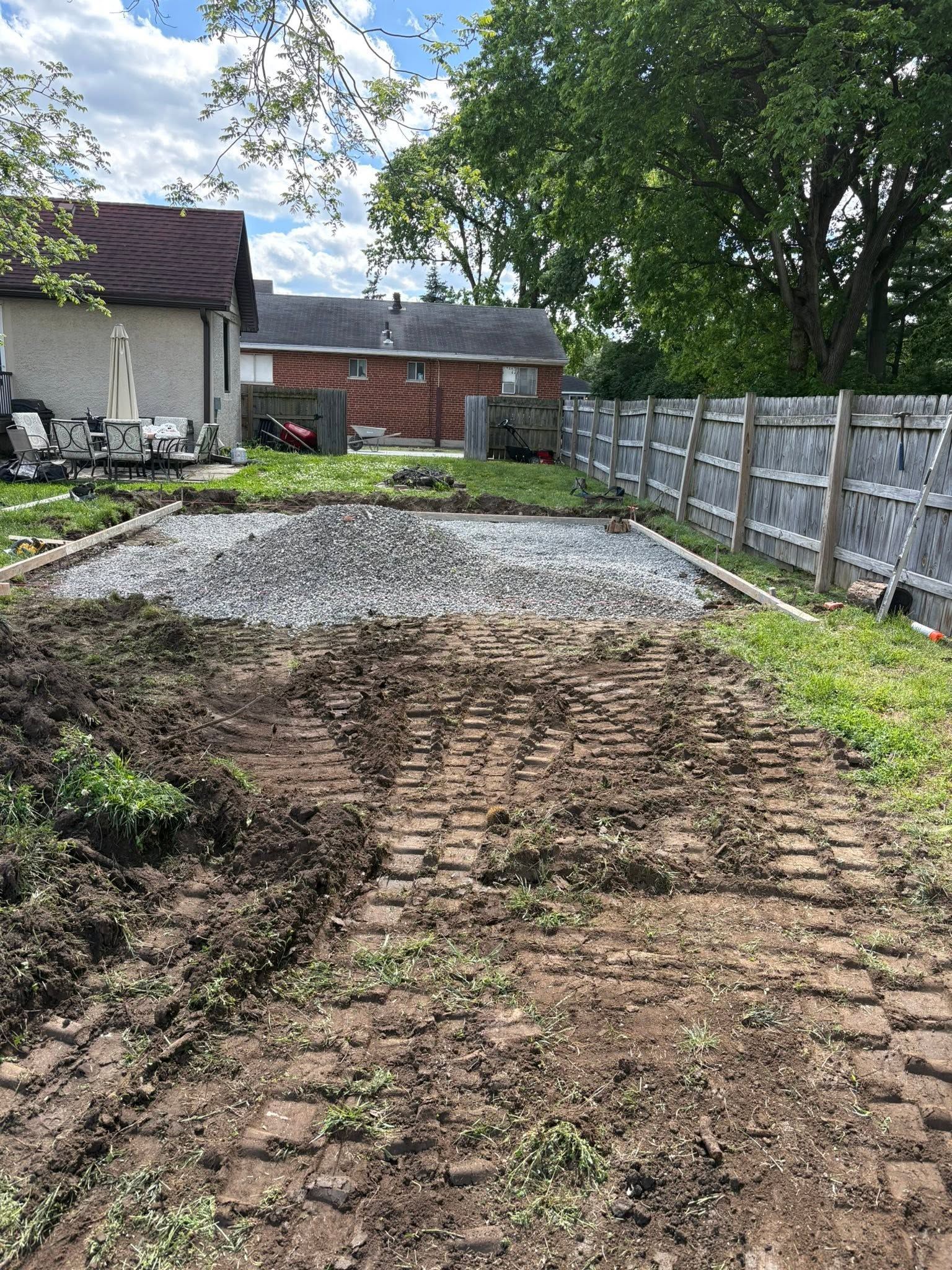 Dirt driveway under construction beside a house, with gravel and tire tracks leading to a wooden fence.