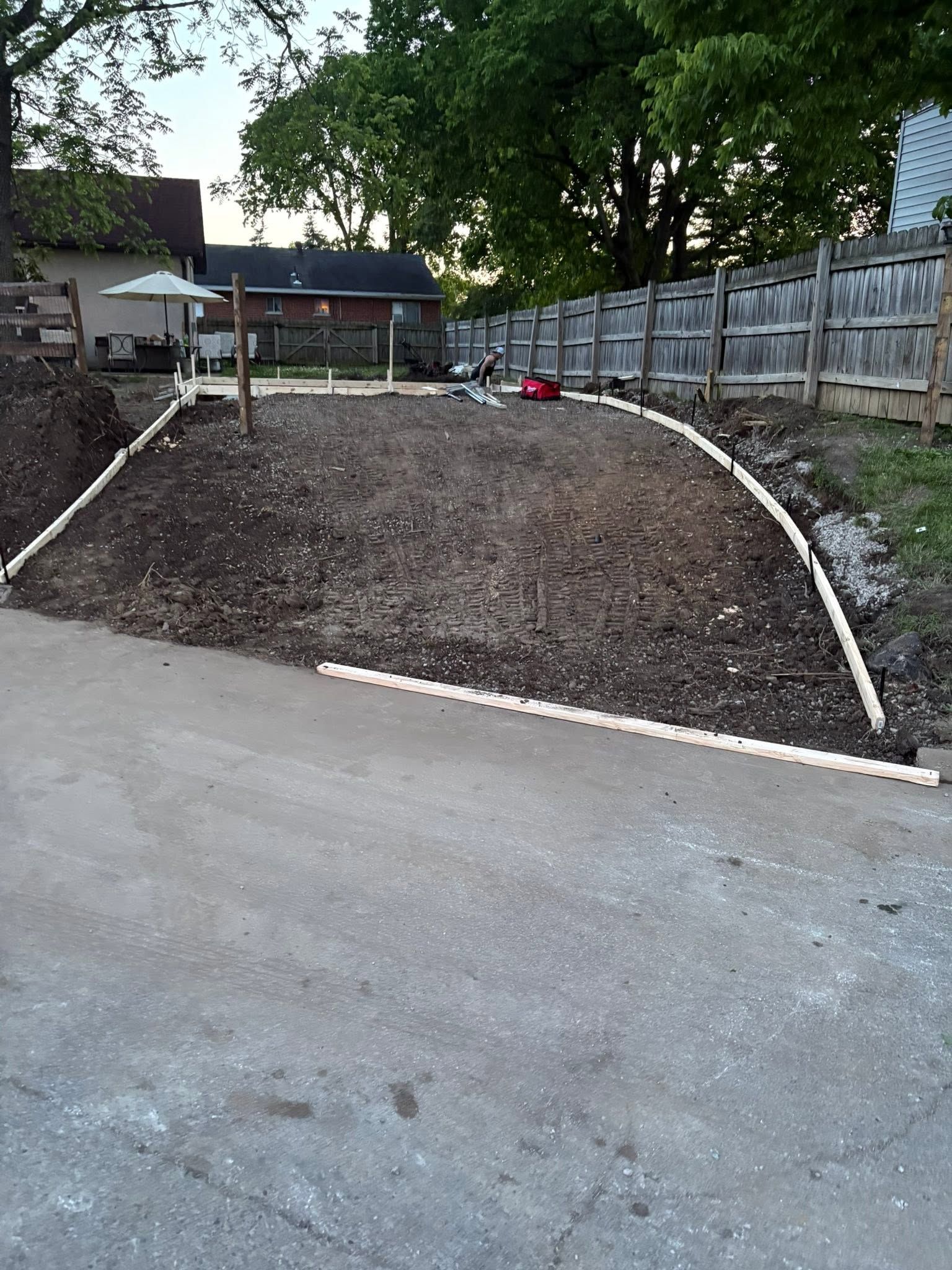 Gravel-covered driveway slope bordered by white pipes, beside a wooden fence and trees in a residential yard