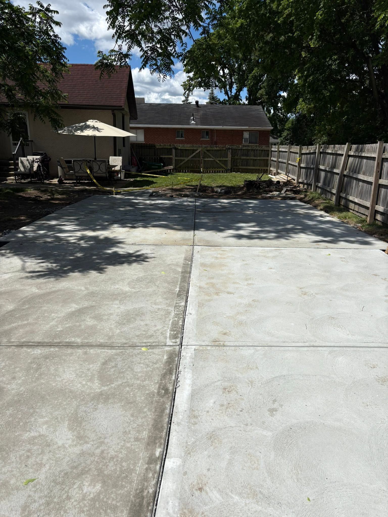 Concrete driveway leading to a house with trees, stone wall, and patio umbrella on a sunny day