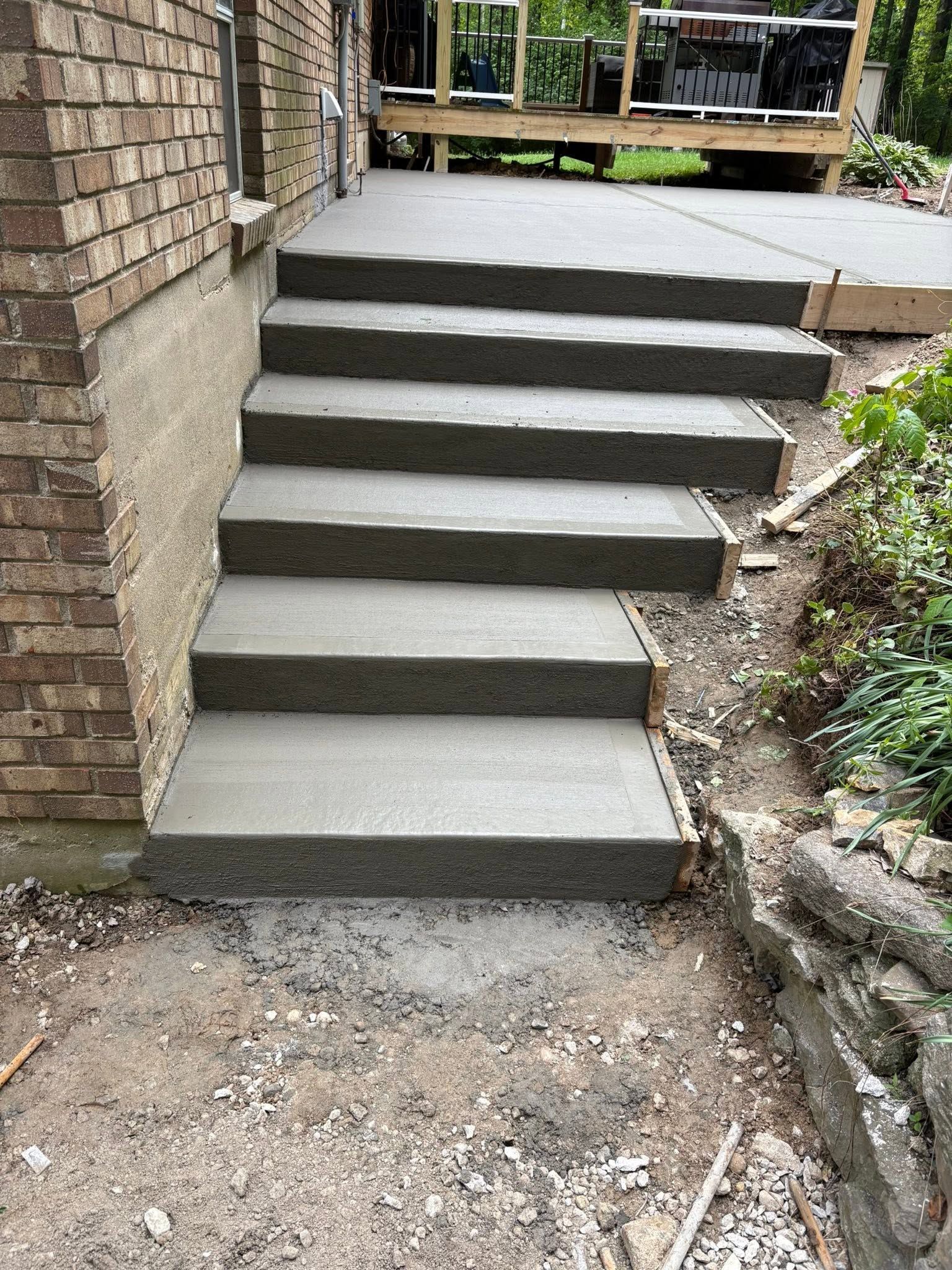 Concrete steps leading from a dirt path to a raised patio beside a brick wall and garden bed