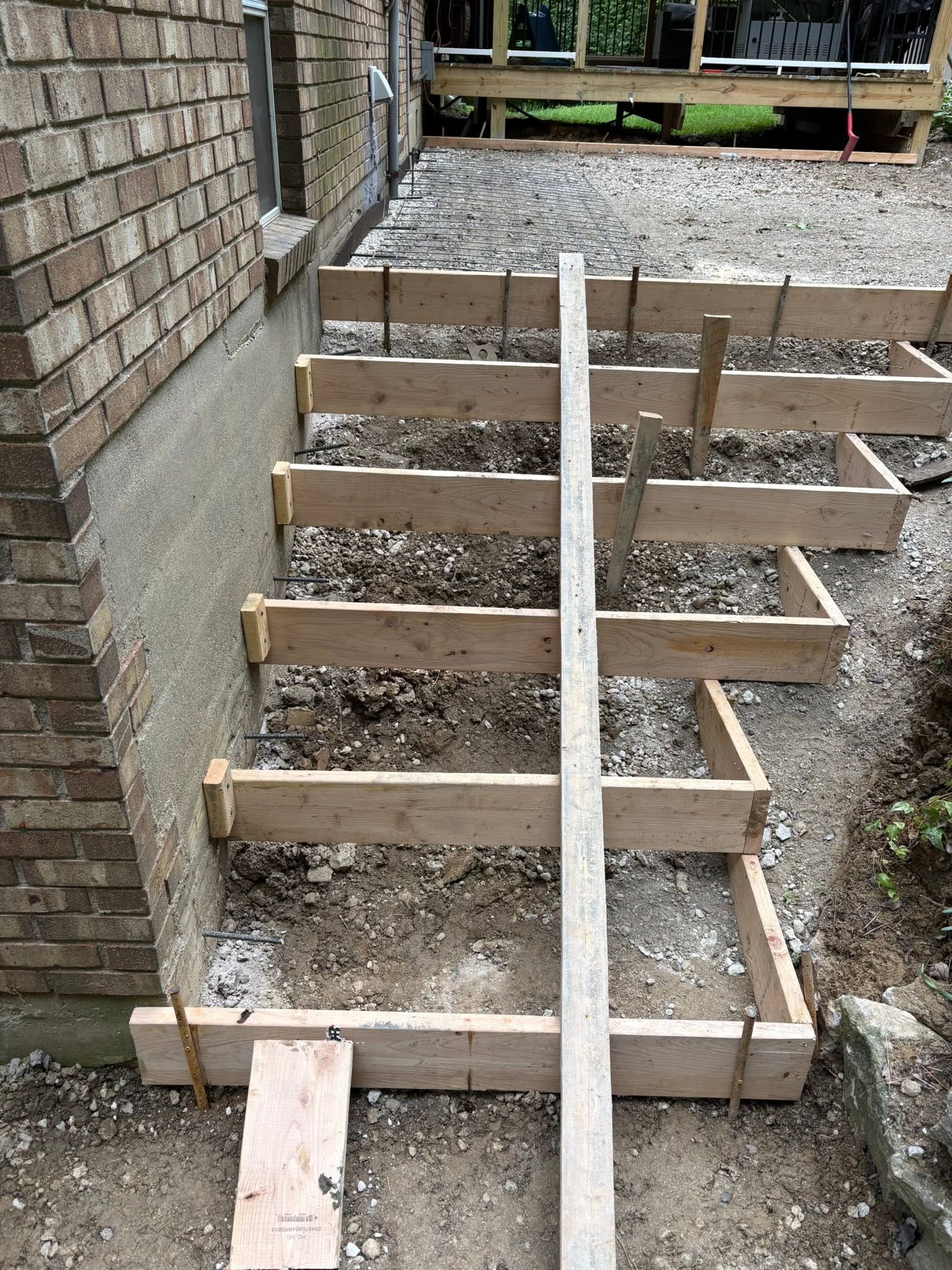 Wooden stair frame under construction beside a brick wall, with gravel ground and support posts.