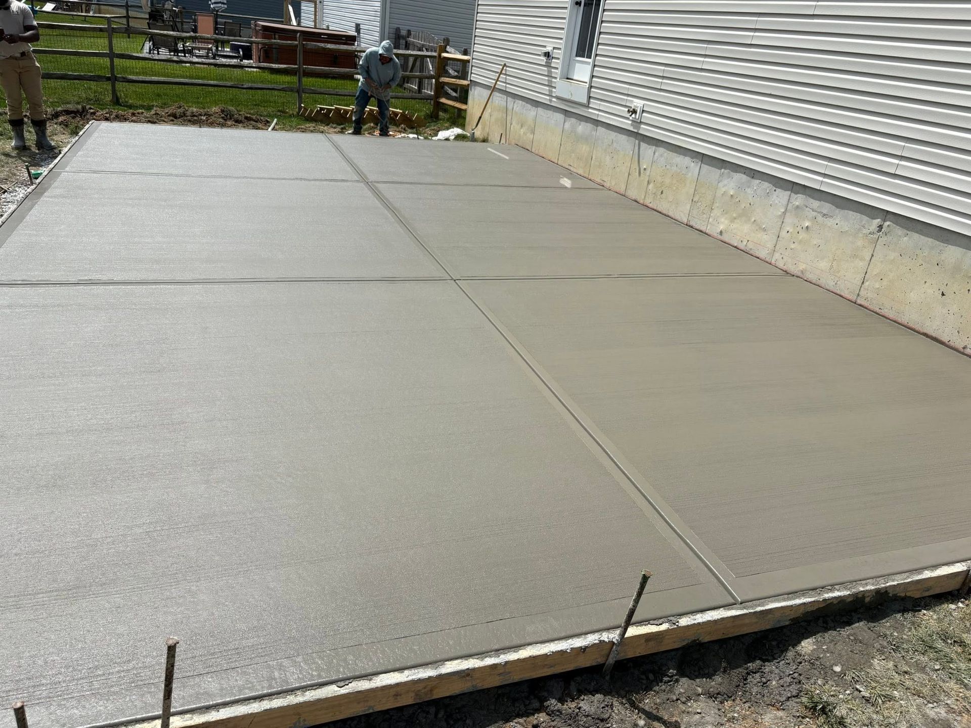 Freshly poured concrete slab beside a house, with a wooden fence and yard in the background