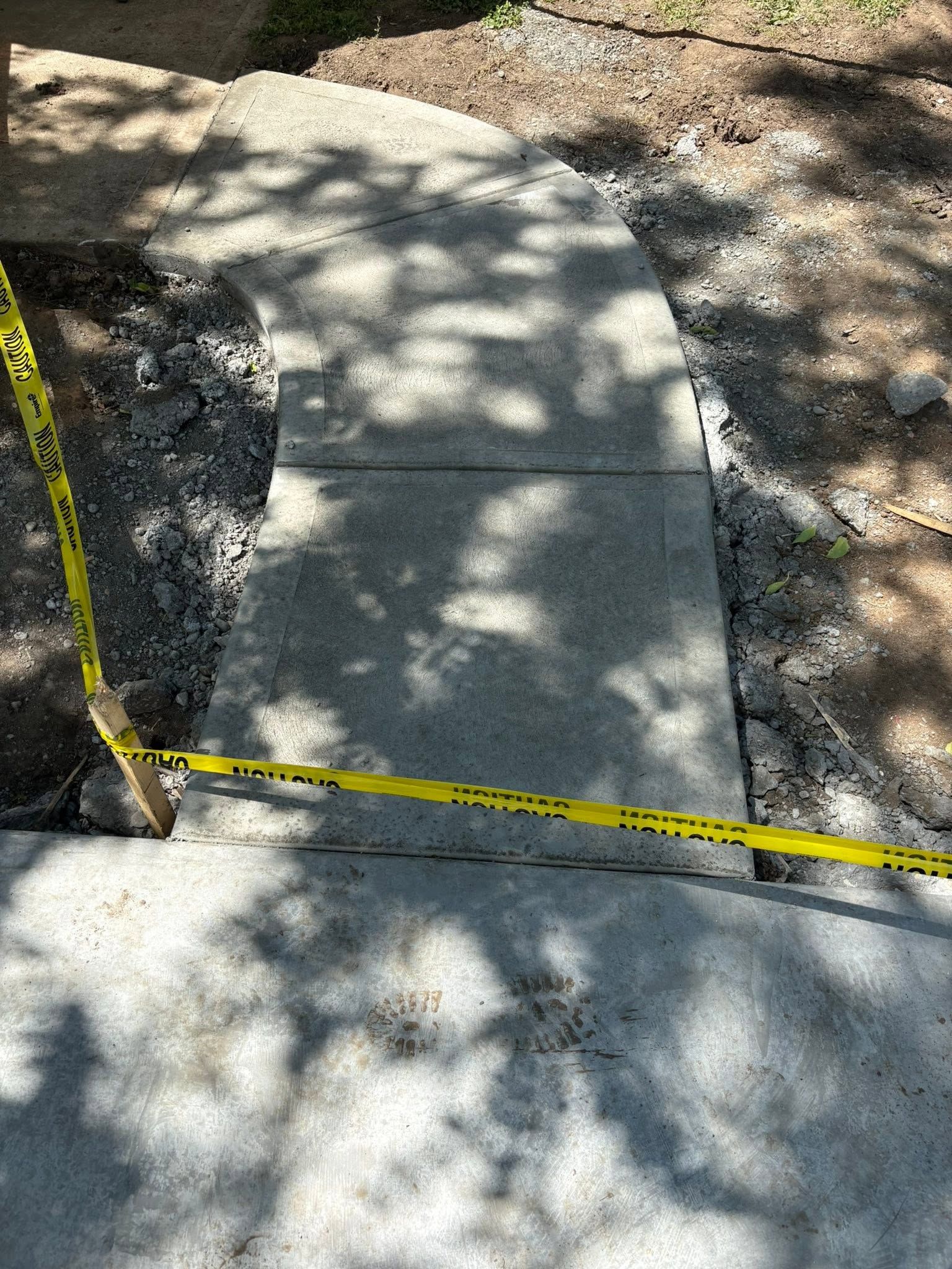 Concrete sidewalk repair area marked with yellow caution tape in a shaded outdoor scene