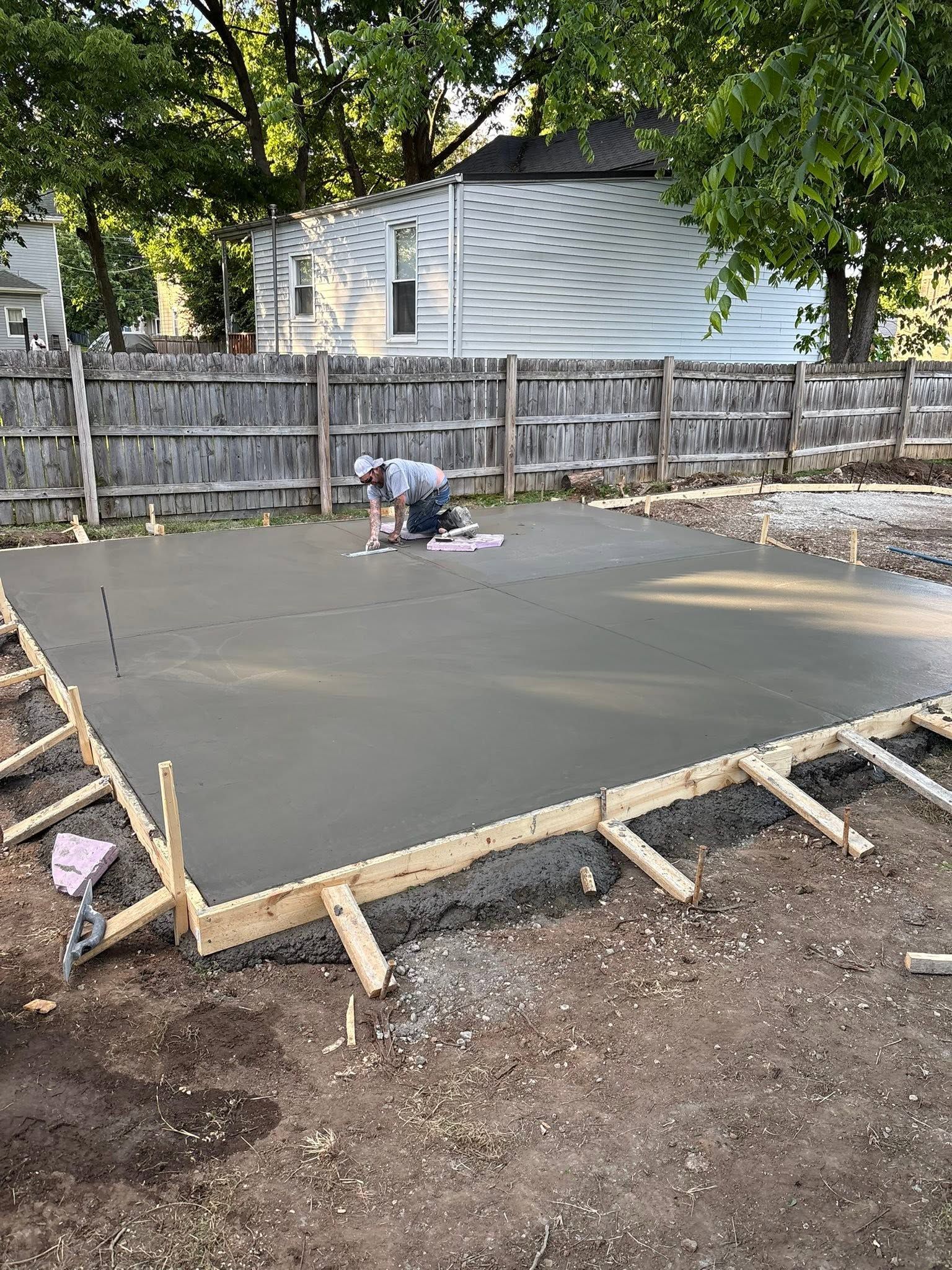 Freshly poured concrete slab in a fenced backyard, with a worker smoothing the surface.